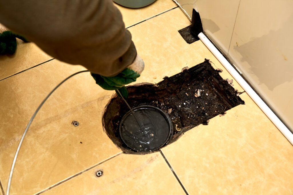 A plumber uses sewer snake to clean blockage in drain on floor.